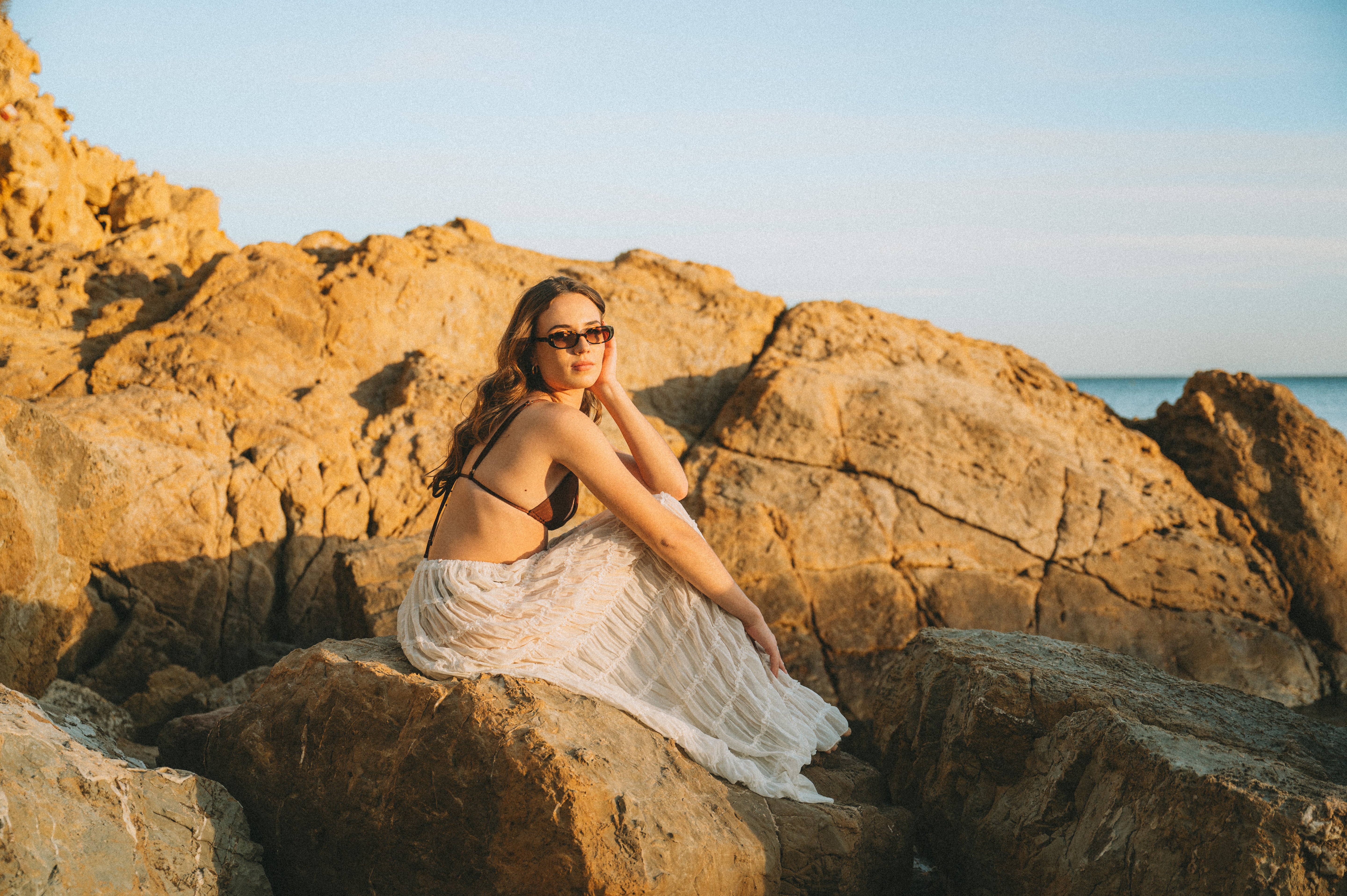 Golden-hour beach portrait of a woman on beach rocks wearing Isla Sunwear tortoise oval sunglasses, designed for smaller faces.