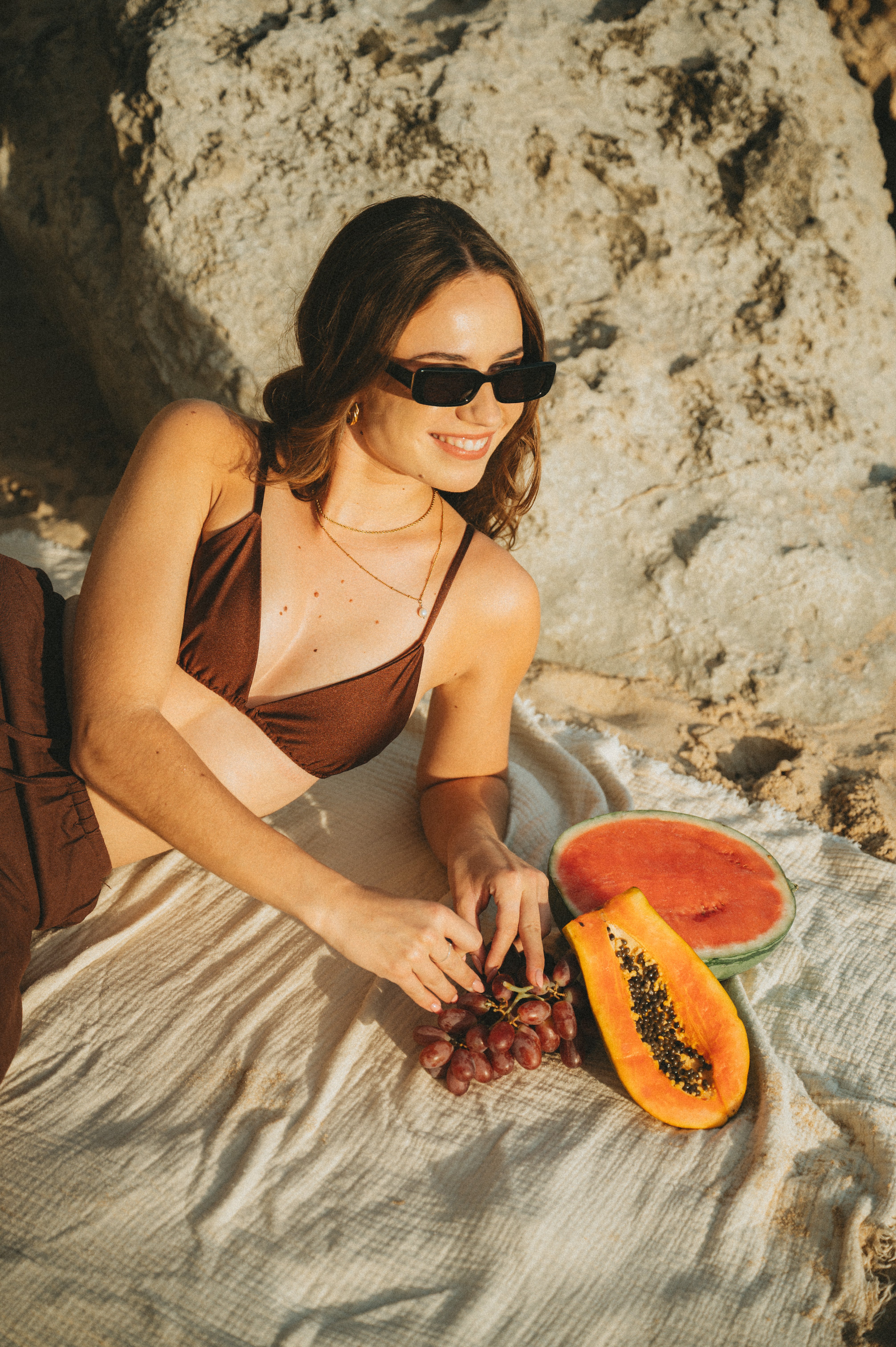 Golden-hour beach portrait of a woman on a beach towel wearing Isla Sunwear black rectangular sunglasses, designed for smaller faces.