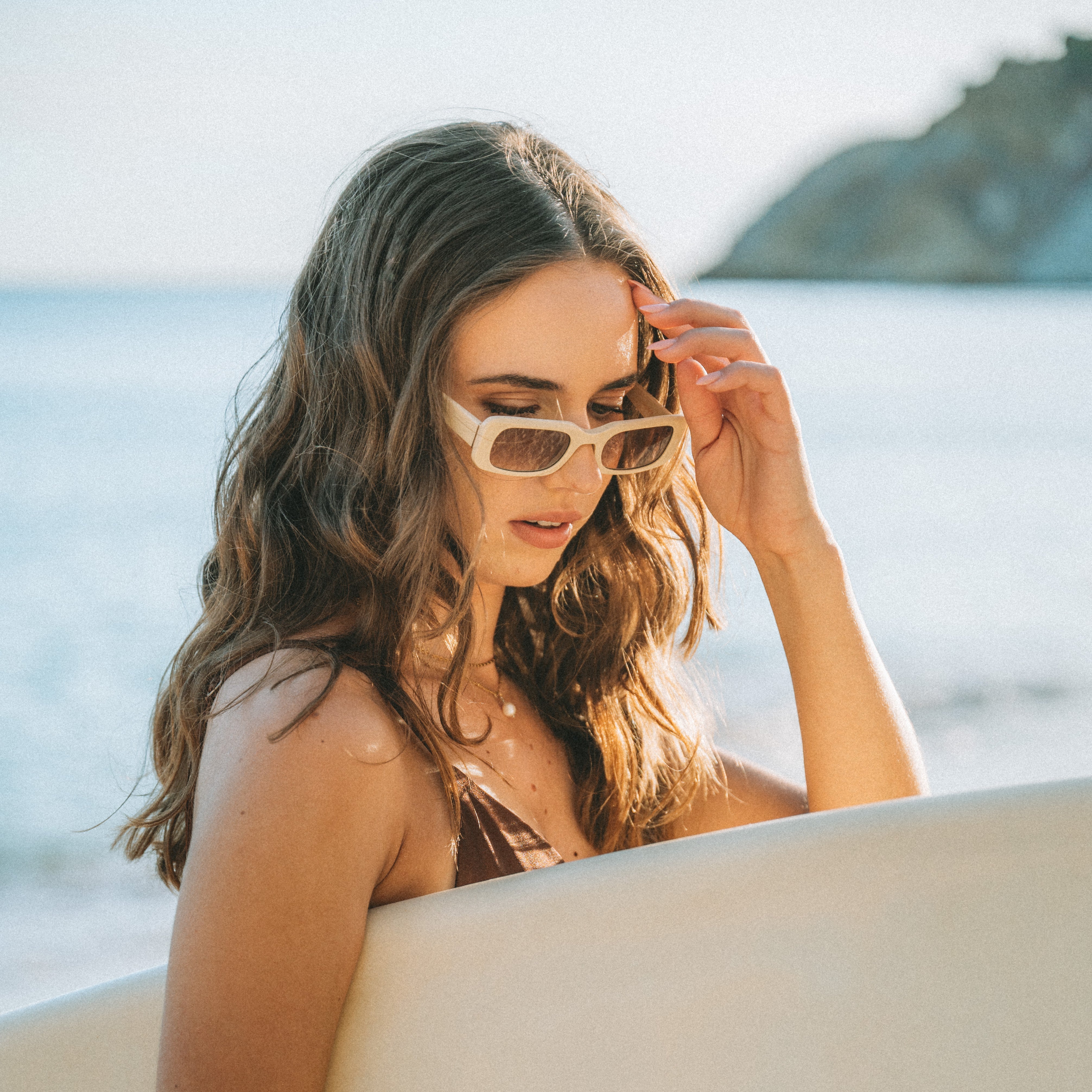 Portrait of a woman holding a surfboard and wearing Isla Sunwear sand rectangular sunglasses, designed for smaller faces.